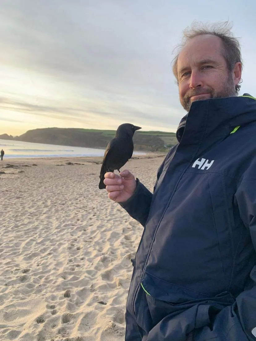 Jon on a Cornish beach at sunrise, a jackdaw perched on his outstretched hand.